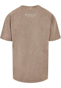 Camiseta marrón de gran tamaño hecha de un tejido suave. Presenta un acabado texturizado y texto impreso en blanco en la parte posterior: "Ámate a ti mismo primero" en árabe e inglés.