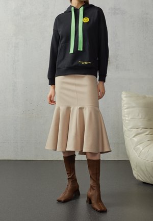 Femme portant un sweat à capuche noir avec cordons verts et un patch smiley jaune, jupe midi évasée beige, et bottes marron texturées mi-mollet.