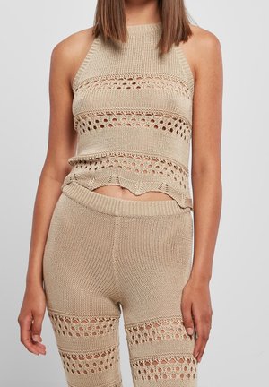 Žena nosící béžový pletený top bez rukávů a odpovídající kraťasy s vysokým pasem, s otevřeným pleteným vzorem na vrchu a stehnech.