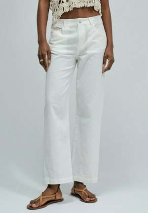 Mujer con pantalones blancos de tiro alto y pernera ancha, sandalias planas beige con tiras y un top beige de crochet, de pie frente a un fondo liso.