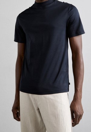 Homme portant un t-shirt noir uni à col roulé et manches courtes, ainsi qu'un pantalon beige clair, sur un fond neutre.