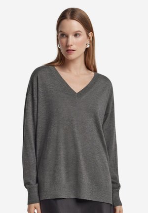 Femme aux cheveux lisses châtain clair portant des boucles d'oreilles créoles en argent et un pull gris décontracté à col en V, regardant légèrement vers la droite.