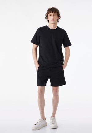 Joven con cabello rizado que lleva camiseta negra y pantalones cortos, zapatillas blancas, de pie con las manos en los bolsillos sobre fondo blanco.