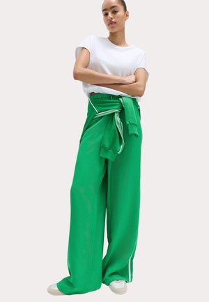 Frau steht mit verschränkten Armen, trägt ein weißes T-Shirt, grüne weite Hosen, einen grünen Pullover, der um die Taille gebunden ist, und weiße Turnschuhe.