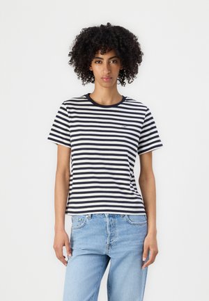 Donna con capelli ricci che indossa una maglietta a maniche corte a righe bianche e nere e jeans azzurri, in piedi davanti a uno sfondo bianco.