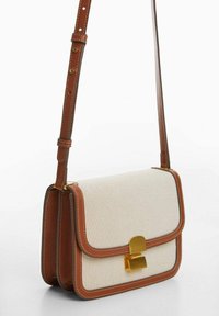 Beige canvas crossbody bag with brown leather accents, gold hardware, and a structured shape. Includes an adjustable strap and stitched details.