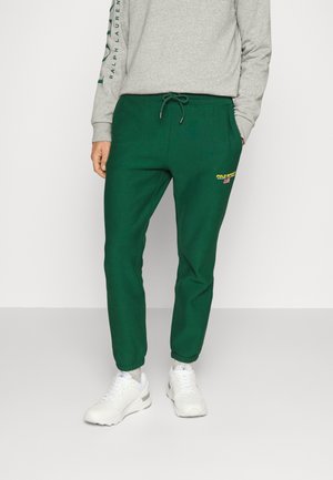 Person, die grüne Polo Sport Jogginghosen mit Kordelzugbund und weißen Sneakers trägt, steht vor einem einfarbigen hellen Hintergrund.