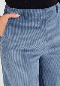 Pantalons en velours côtelé bleu avec texture côtelée verticale, taille ajustée et poches latérales. Le tissu a une apparence douce et veloutée.
