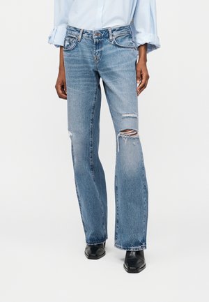 Άτομο που φοράει ανοιχτό μπλε σκισμένο τζιν με ίσια γραμμή, μαύρα μυτερά παπούτσια και ανοιχτό μπλε πουκάμισο με μανίκι γυρισμένο προς τα πάνω.