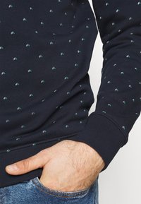 Suéter de rayas en azul marino con pequeños diseños geométricos en verde y blanco. Presenta puños y dobladillo acanalados. El material parece texturizado y de peso medio.