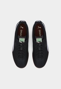 Ζευγάρι μαύρα παπούτσια Puma με λευκές πλαϊνές ρίγες και πράσινες ετικέτες με το λογότυπο, εμφανιζόμενα από πάνω σε απλό φόντο.