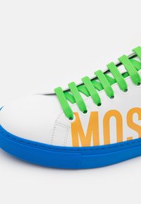 Białe sneakersy z wyraźnym pomarańczowym napisem "M.O.S", zielonym teksturowanym sznurówkami i niebieską gumową podeszwą. Gładka skórzana powierzchnia i okrągły design noska.