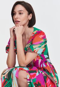 Kolorowa sukienka o krótkich, bufiastych rękawach. Posiada odważne, abstrakcyjne wzory w kolorach zielonym, różowym, czerwonym i niebieskim. Miękka tekstura materiału.