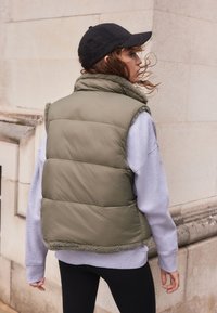 Vesta verde căptușită cu un colier pufoasă, purtată peste un hanorac gri. Vesta are un exterior neted și un design scurt cu o linie de tiv.