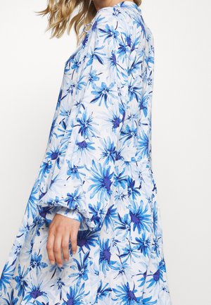 Nainen, jolla on päällään löysä, pitkähihoinen mekko, jossa on sinivalkoinen kukkakuvio, seisoo yksinkertaisen valkoisen taustan edessä.