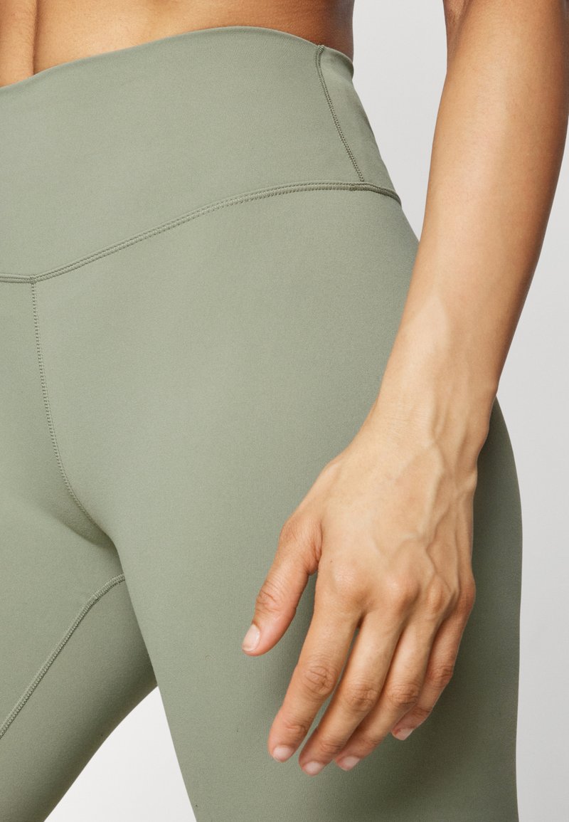 Hoog getailleerde, olijfgroene legging gemaakt van een gladde, elastische stof met platte naden en een aansluitend ontwerp, waarbij een hand op de taille is geplaatst.