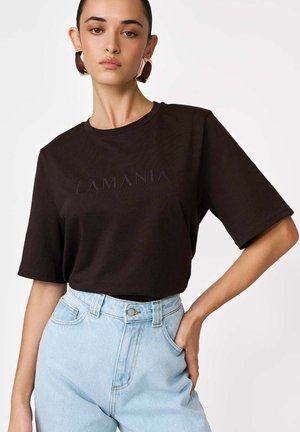 Kobieta w czarnym, oversize T-shircie z napisem "LAMANIA", jasnoniebieskich, wysokim stanem jeansach i dużych brązowych kolczykach, pozująca z ręką w kieszeni.