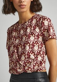 Top de manga corta, con un patrón rojo y un frente anudado, que presenta formas de diamante y geométricas en tonos crema y rosa, hecho de una tela suave.