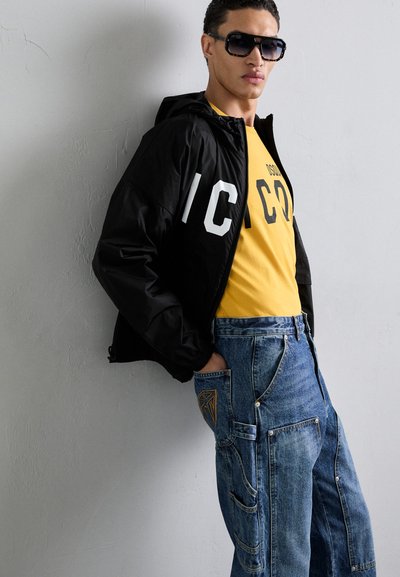 Veste zippée noire avec des lettres blanches, t-shirt jaune avec un motif, et jeans en denim bleu avec des coutures détaillées et plusieurs poches.