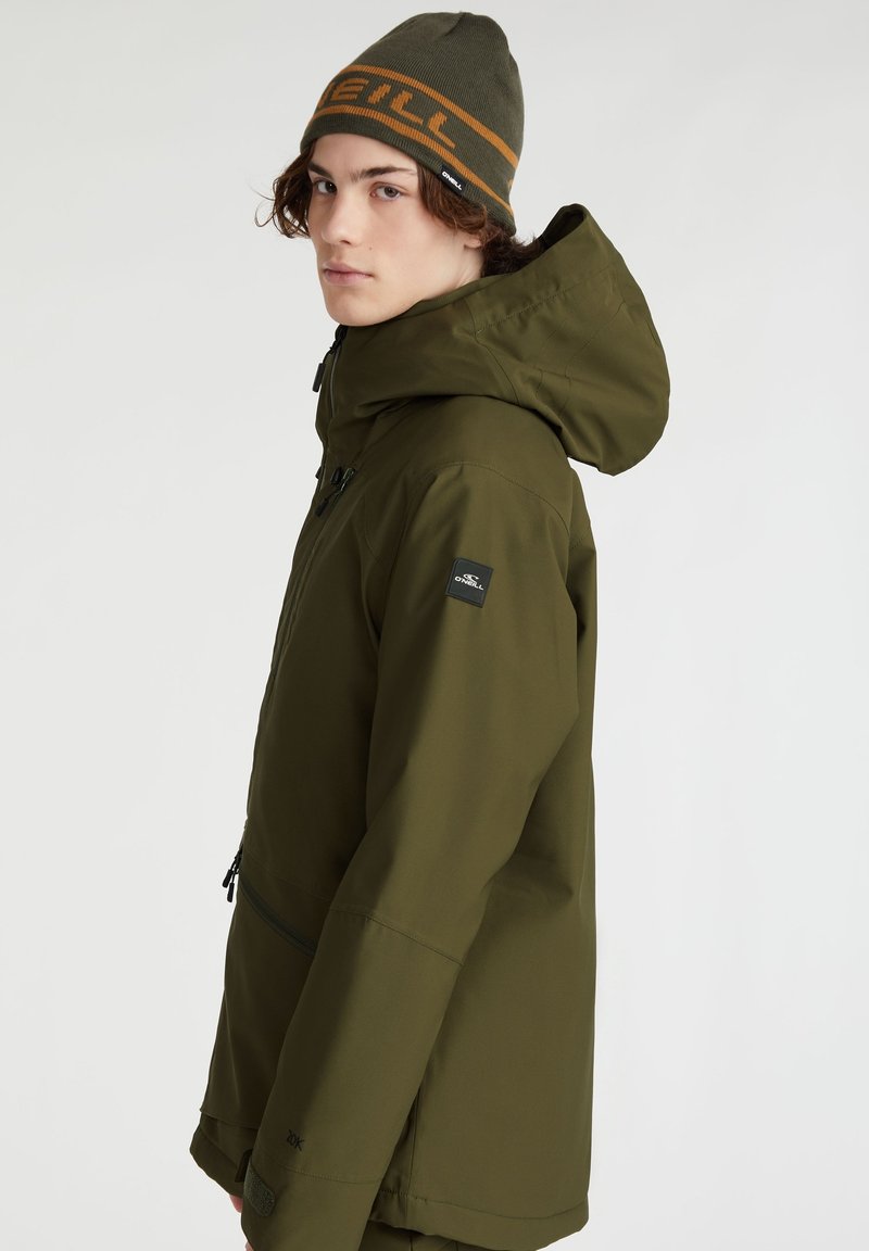 Olivgrüne wasserdichte Jacke mit Kapuze, mit Reißverschlusstaschen und einem Logo-Patch am Ärmel. Getragen mit einer passenden Mütze.