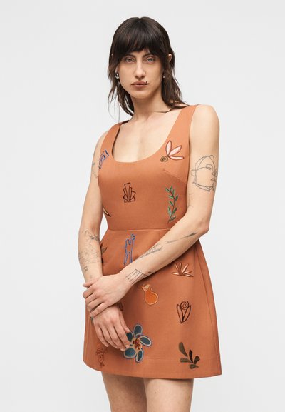 Γυναίκα με σκουρόχρωμα μαλλιά και τατουάζ, που φοράει αμάνικο φορεματί απόχρωσης σκουριάς με πολύχρωμα, κεντημένα αφηρημένα και λουλουδάτα σχέδια, στέκεται μπροστά σε αplain φόντο.