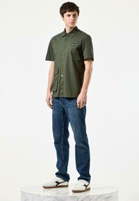 Groene overhemd met korte mouwen, een kraag, een knoopsluiting aan de voorkant en een zak; gecombineerd met blauwe spijkerbroek en witte sneakers met zwarte accenten.