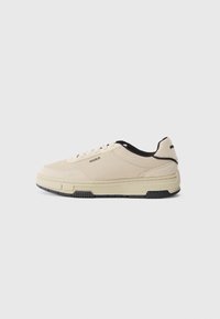 Beige sneakers med teksturert stoffoverdel, svarte detaljer på kragen, en tykk hvit såle og en subtil merkevarelogo på siden.