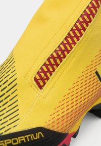 Zapato de escalada amarillo con acabado texturizado, que presenta una cremallera roja y un patrón geométrico. La suela negra y los acentos amarillos realzan el diseño.