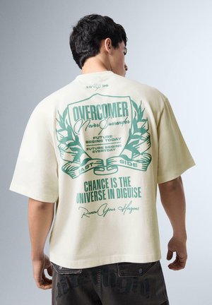 Man die een crèmekleurig oversized t-shirt draagt met groene tekst en lauwerkransontwerp met de tekst "Overcomer Never Surrender, Chance is the Universe in Disguise."