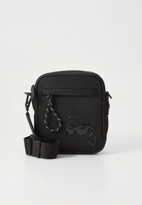 Juoda per petį nešiojama rankinė su lygiu paviršiumi, priekyje esančiu užtrauktuko kišene ir iškilusiu akinukų dizainu. Komplekte nuimamas dirželis.