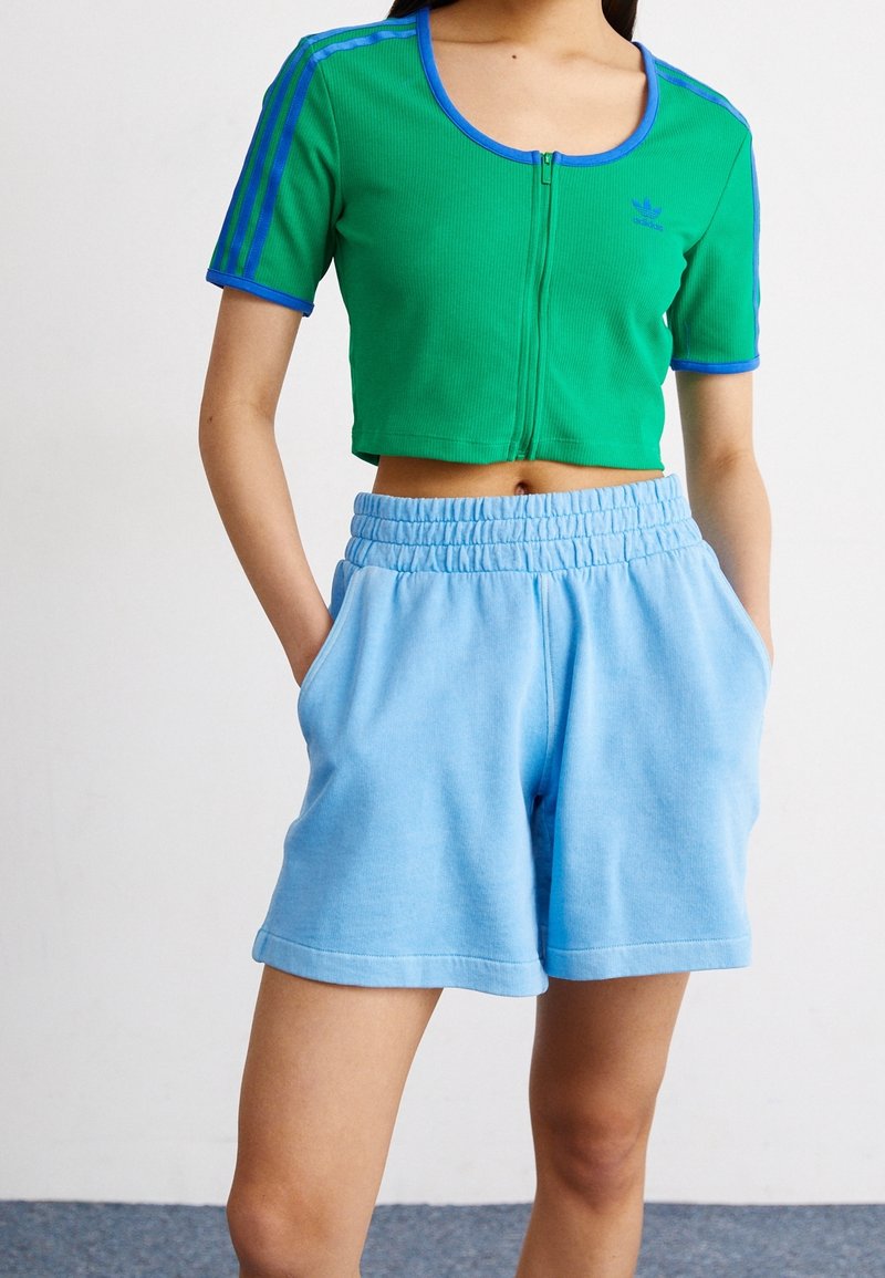 Zelený pruhovaný top s modrým lemováním a zipem, kombinovaný se světle modrými šortkami na šňůrku se bočními kapsami.