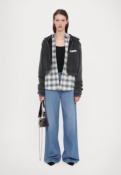 Dunkelgrauer, verkürzter Hoodie über einem schwarzen Tanktop, karierter Hemd und weit geschnittenen blauen Jeans. Als Accessoire dient eine kleine glänzende Handtasche.