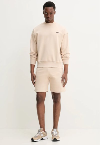 Completo felpa e pantaloni corti beige realizzato in tessuto morbido. La felpa ha un collo rotondo, polsini a coste e dettagli del logo. Sneakers in tonalità neutre con accenti.