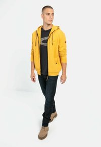 Gul zip-up hoodie med snoretræk, båret over en sort t-shirt. Mørkeblå jeans og lysebrune sko fuldender outfittet.