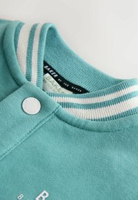 Zelená mikina s žebrovaným límcem s bílými pruhy. Obsahuje knoflík s logem a štítek označený "BAKER by TED BAKER."