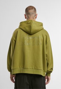 Olīvu zaļa pārnesuma kapuce ar teksturētu audumu, kuras aizmugurē ir sudraba rhinestone apdruka, kas vēsta "Niekur nav tagad šeit."
