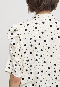 Blusa blanca con lunares negros, con mangas abullonadas y cuello con solapa. Textura de tela suave con acentos de patrón distribuidos de manera uniforme.