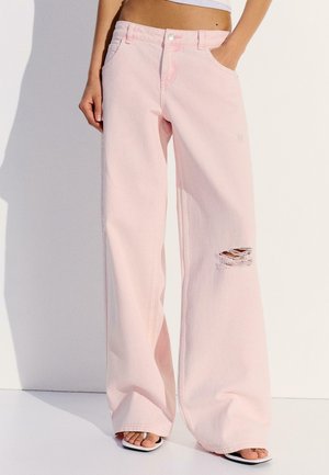 Frau trägt hoch taillierte, weit geschnittene hellrosa Jeans mit einem Riss am Knie und steckt die Hand in die Tasche, vor einem schlichten weißen Hintergrund.