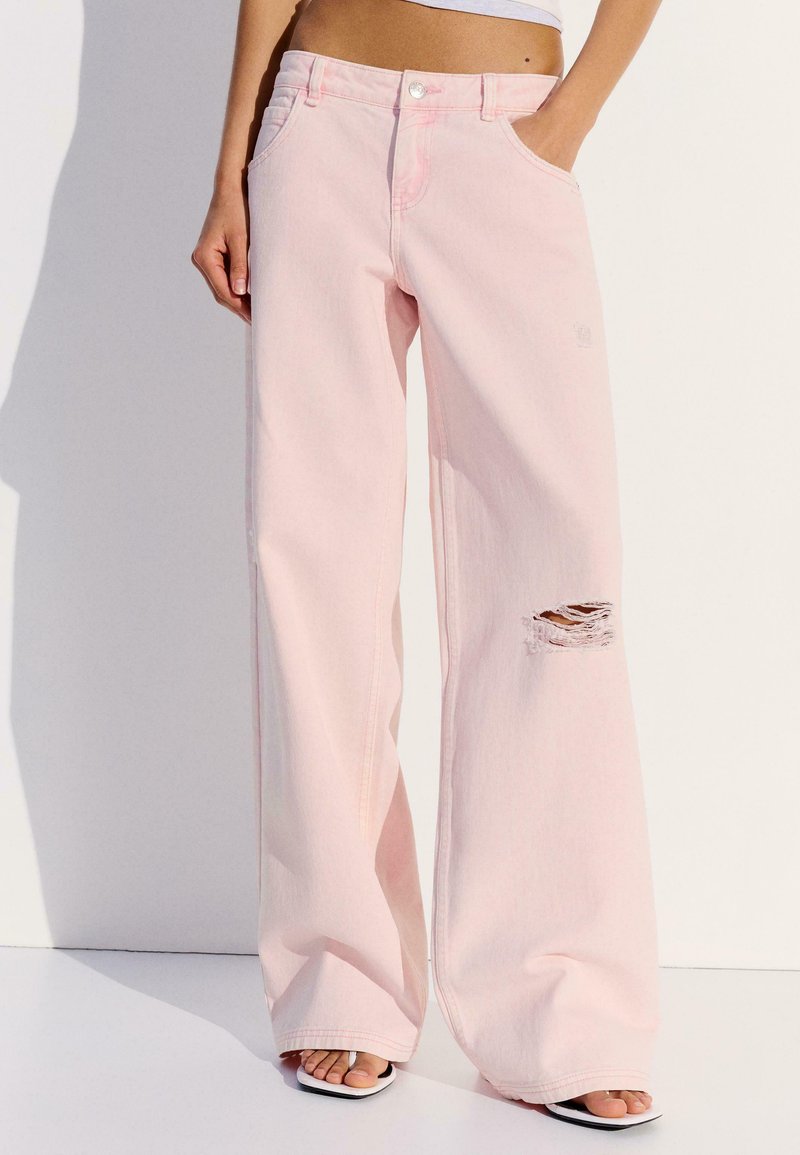 Femme portant un jean rose pâle taille haute, à jambes larges, avec un détail déchiré au genou et la main rentrée dans la poche, sur un fond blanc uni.