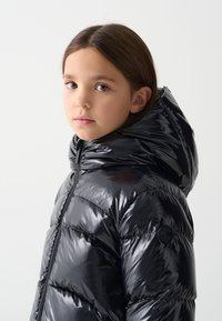 Melnas pūkains jaka ar spīdīgu virsmu, kapuci un rāvējslēdzēju priekšpusē. Ar šūtiem segmentiem un logotipa uzšuvumu piedurknē.