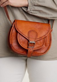 Braune Leder-Crossbody-Tasche mit einer Klappe, Schnallendetails und gestickten Akzenten. Kompakte, runde Form mit glatter Textur.