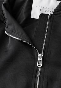 Veste noire avec une finition lisse et mate ; comporte une fermeture éclair en métal sur le devant et une étiquette indiquant "STREET ONE STUDIO."