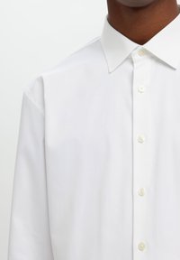 Λευκό πουκάμισο με κουμπιά, διαθέτοντας κλασικό γιακά, λεία υφή και κουμπιά που μοιάζουν με μαργαριτάρια, παρουσιάζεται σε ουδέτερο φόντο.