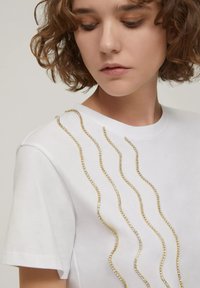 T-shirt bianco in cotone con decorazioni ondulate in oro e cristallo lungo la spalla sinistra. Maniche corte e scollatura rotonda.