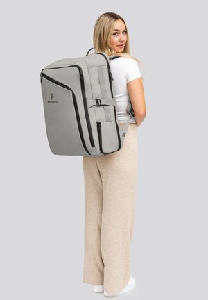 Sac à dos gris avec des zippers et des accents noirs, de forme rectangulaire, avec des bretelles rembourrées, logo sur le devant, porté sur le dos sur un fond gris clair.