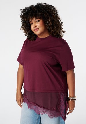 Vrouw met krullend haar die een bordeauxkleurige gelaagde top met kanten zoom en lichtblauwe jeans draagt, staand tegen een effen witte achtergrond.