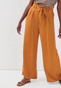 Pantalons à jambes larges de couleur orange vif, fabriqués en tissu doux avec un devant plissé et une taille nouée. Assortis avec des sandales plates marron clair.