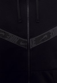 Svart zip-up hoodie med en mjuk textur, dragsko och ett brett horisontellt band med en tonad Nike-logotyp över bröstet.