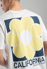 White cotton t-shirt featuring a large yellow flower graphic and "CALIFORNIA" text in blue at the back. Short sleeves, crew neckline.