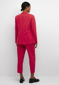 Completo a due pezzi aderente di un vivace rosa con giacca e pantaloni cropped, realizzato in tessuto morbido. Abbinato a scarpe slip-on nere.